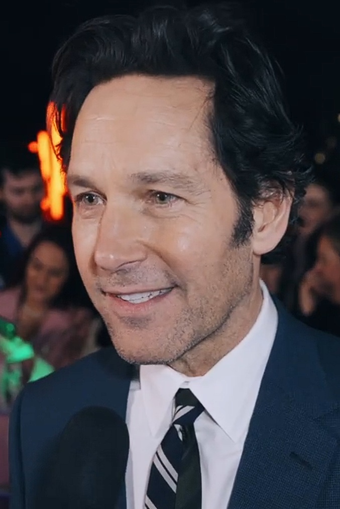 Close-up of a man in a blue suit and striped tie, smiling and speaking into a microphone, with blurred people and lights in the background.