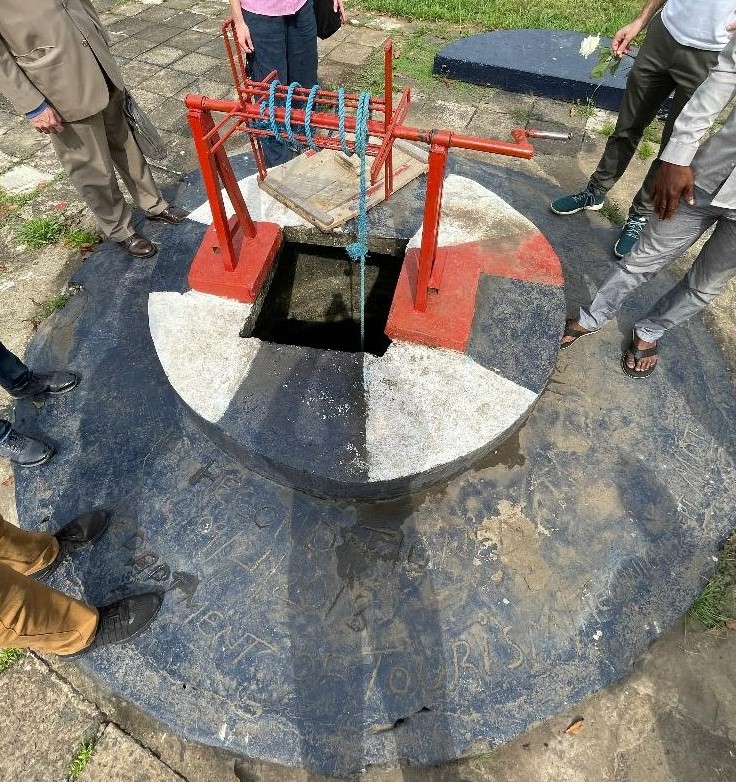 A circular ancient water well with a red manual winch and a blue rope, surrounded by people standing around it on a paved ground.
