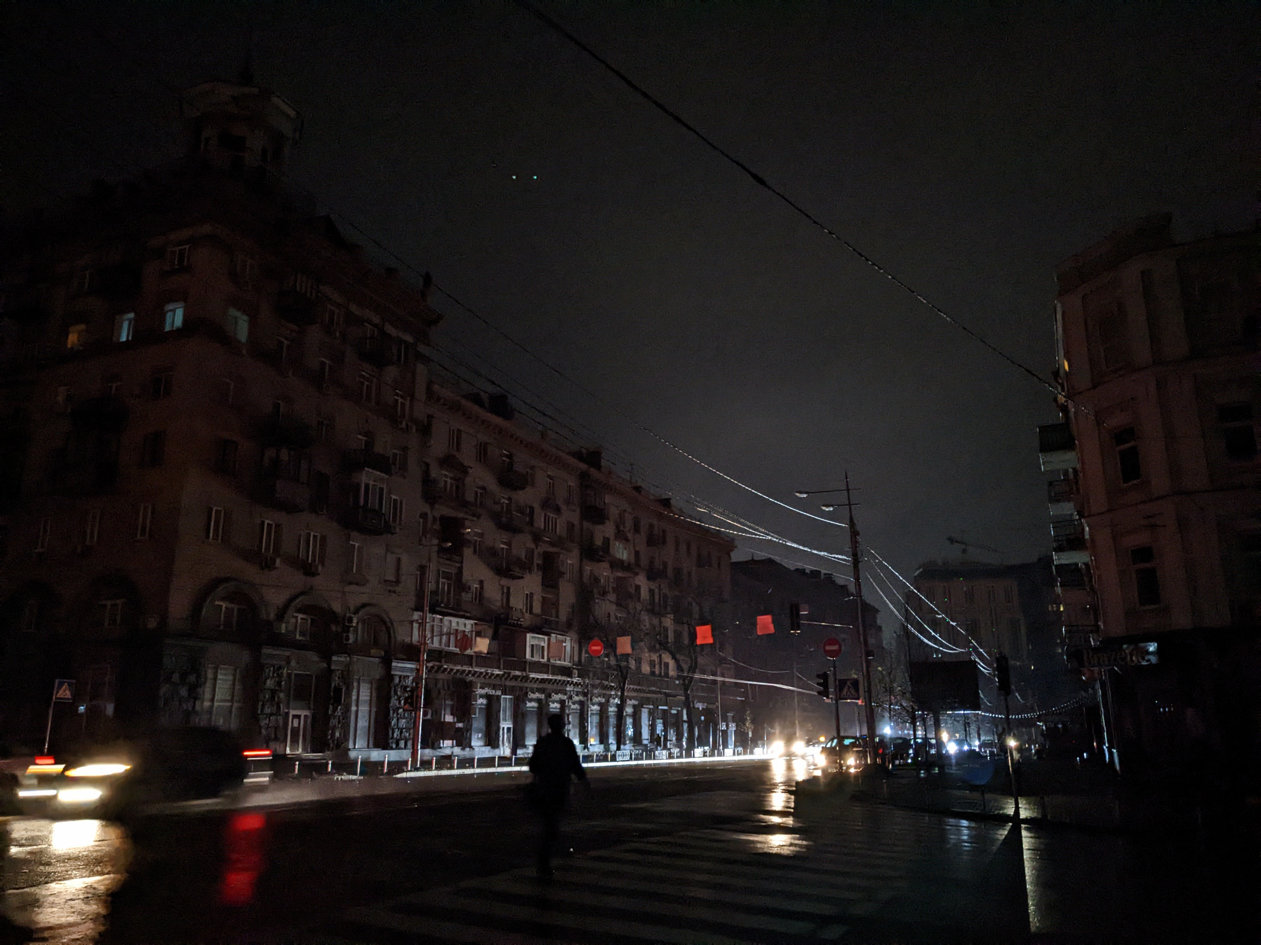 Dark street with buildings on either side, few vehicle headlights and a person crossing in low light