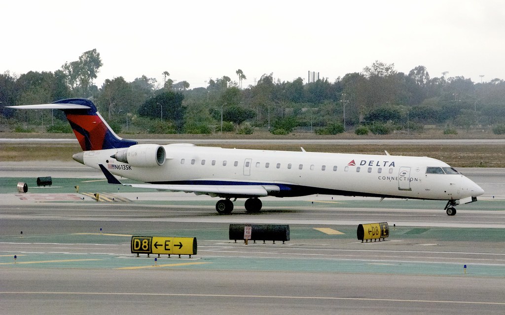 White Delta Connection Bombardier RJ airplane taxiing on runway with trees and infrastructure in background