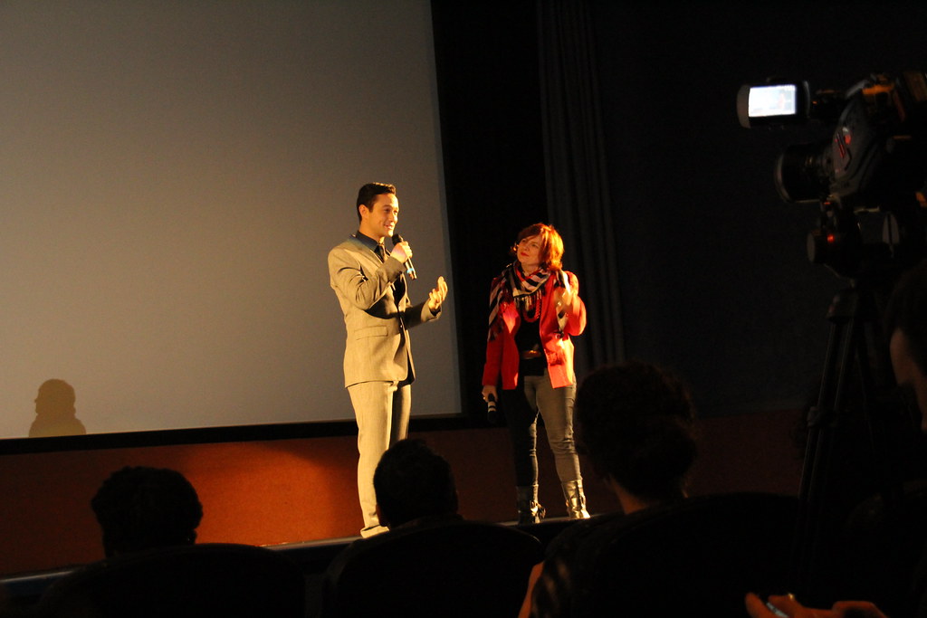 Joseph Gordon-Levitt standing on stage holding a microphone, accompanied by a woman also holding a microphone, with an audience and a camera present.