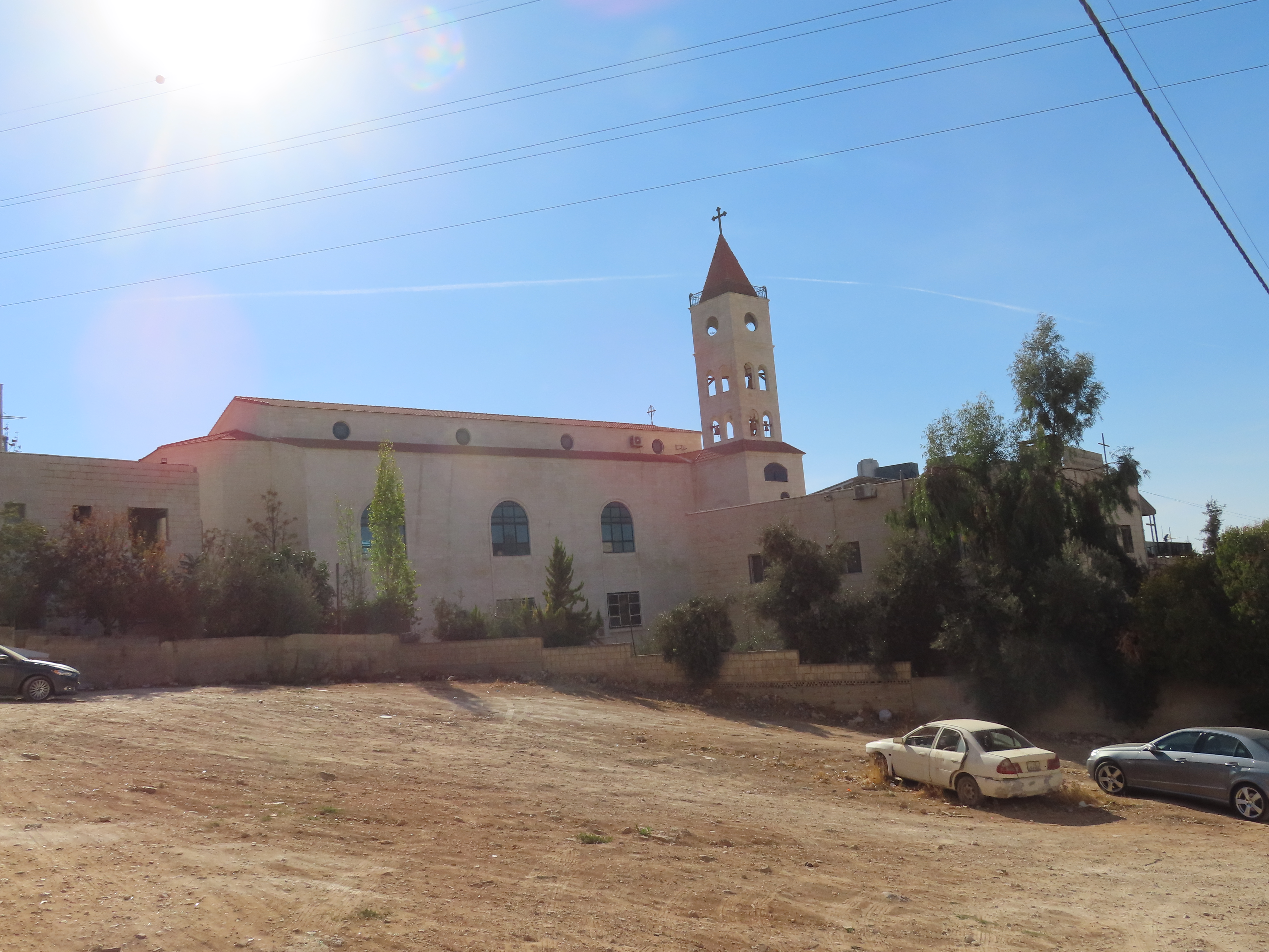 Exterior of a church building with a tall steeple topped with a cross, surrounded by some trees and cars parked on a dusty ground.