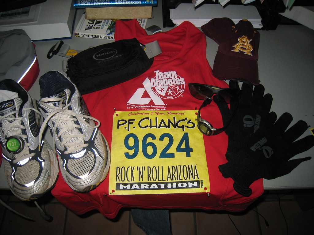 Marathon running gear including shoes, a red sleeveless shirt with a race bib, gloves, sunglasses, a watch, and a cap laid out on a table.