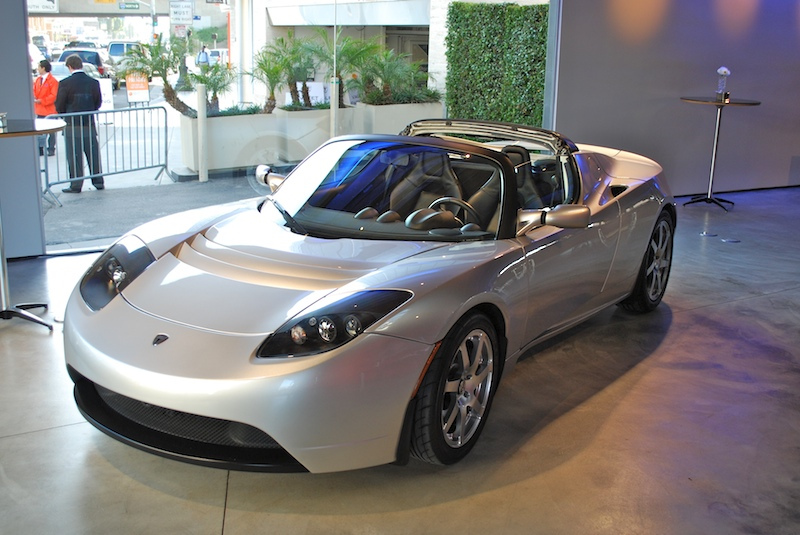 Silver Tesla Roadster electric car parked indoors with a few people and plants visible in the background