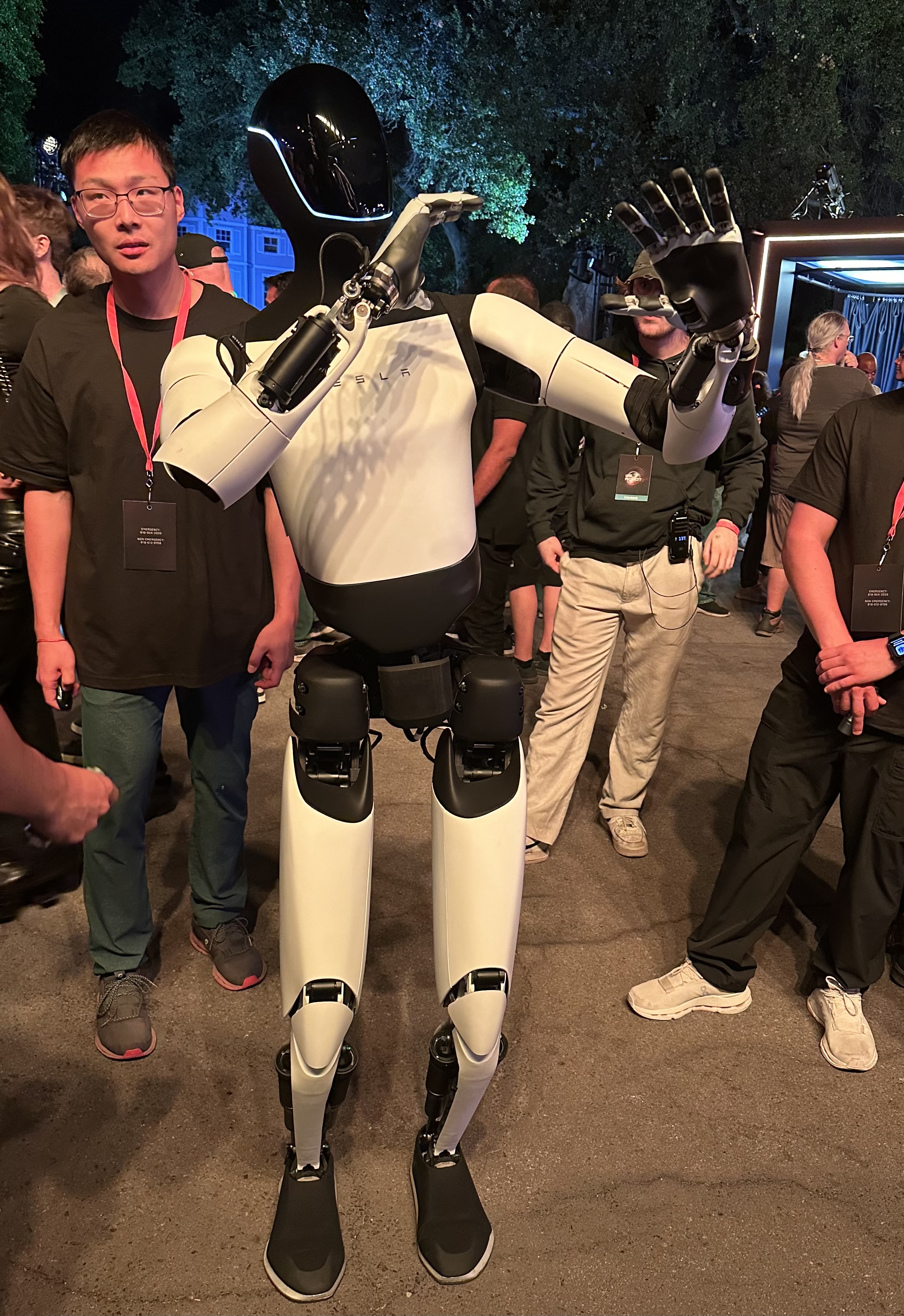 Tesla humanoid robot standing outdoors with people around at night.