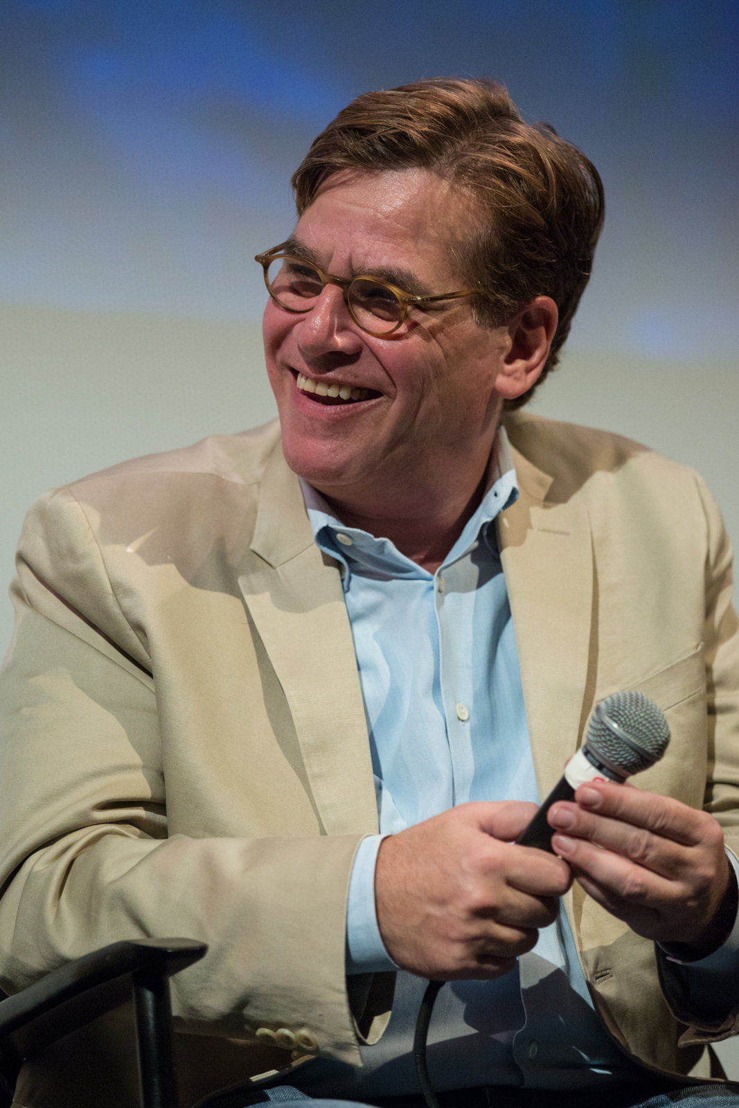 Man wearing glasses, beige jacket and light blue shirt holding a microphone and smiling