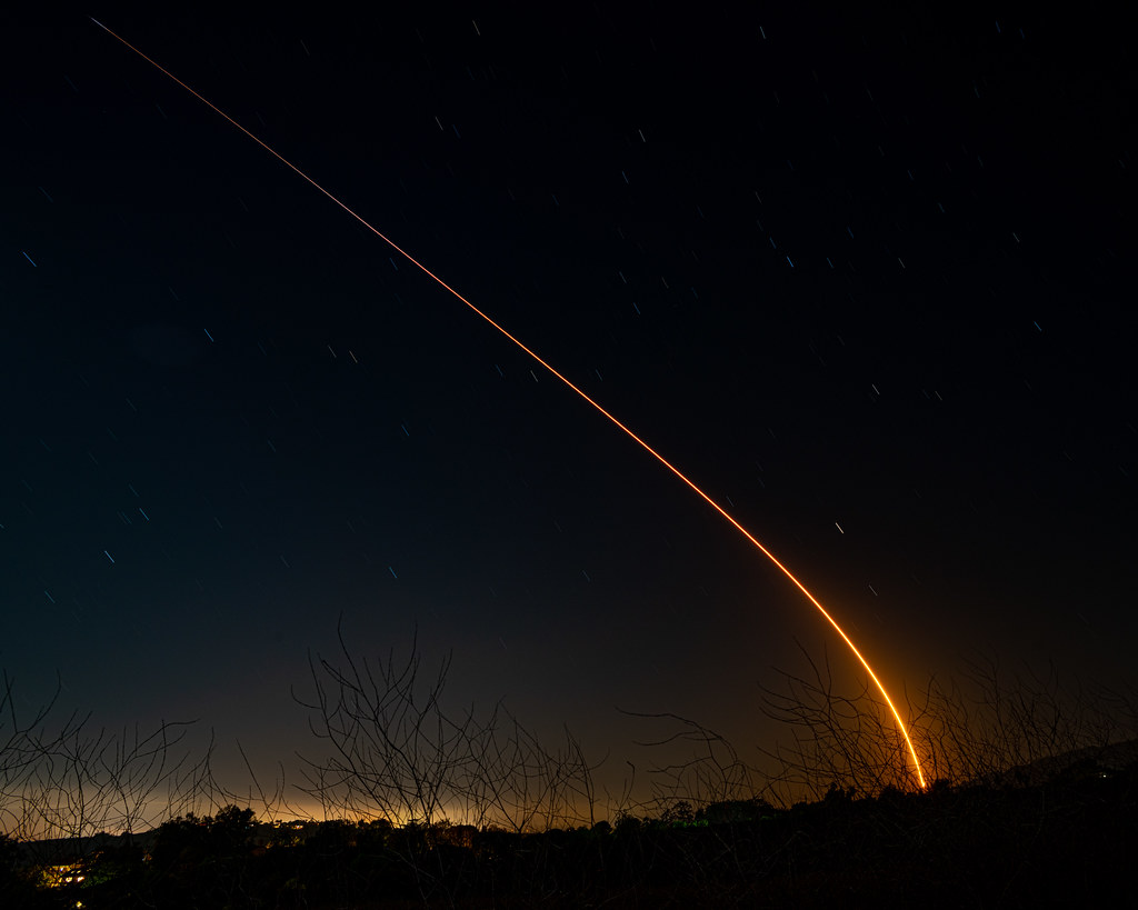 Night sky with a curved bright orange trail of a rocket launch rising from behind hills with trees and branches in the foreground.