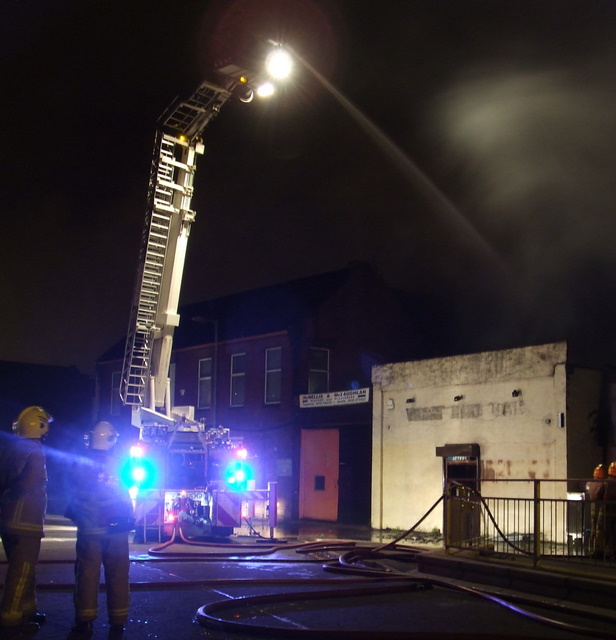 Firefighters using a ladder truck to spray water on a building at night, with hoses on the ground and light from the fire truck illuminating the scene.